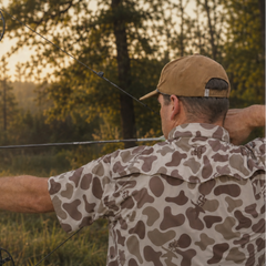 A man in a camouflage shirt and tan cap draws a compound bow outdoors in a wooded setting during golden hour.